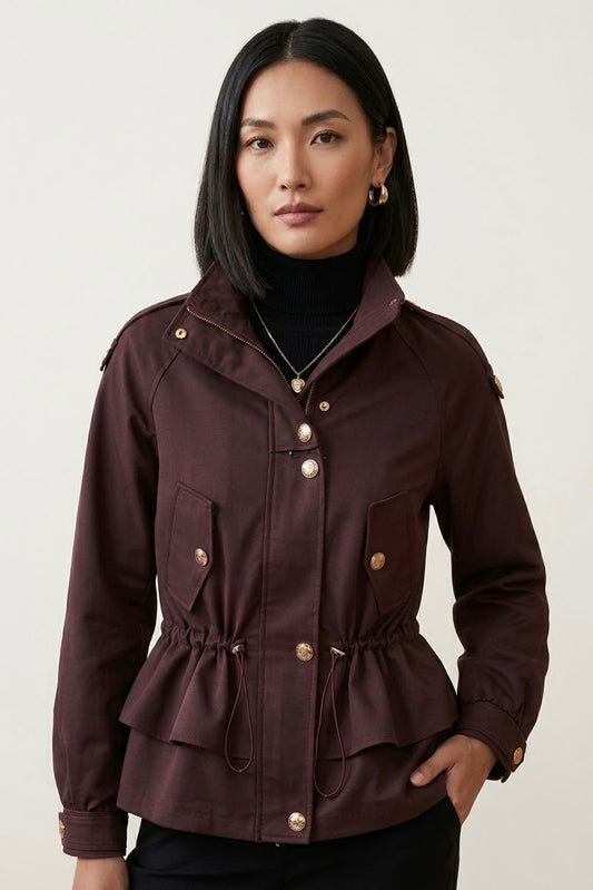 A woman modeling a dark burgundy peplum jacket with gold-tone buttons and a drawstring waist, worn over a black turtleneck against a light gray studio background.