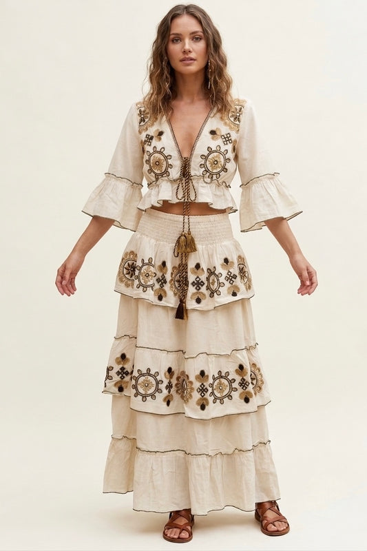 A woman modeling a cream-colored bohemian-style two-piece set featuring a cropped jacket with bell sleeves and a matching tiered maxi skirt, both decorated with intricate brown and gold geometric embroidery.
