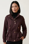 A woman modeling a dark burgundy peplum jacket with gold-tone buttons and a drawstring waist, worn over a black turtleneck against a light gray studio background.