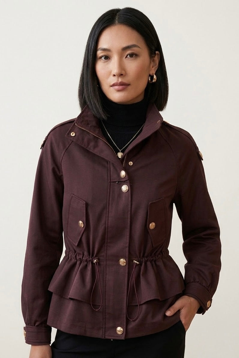A woman modeling a dark burgundy peplum jacket with gold-tone buttons and a drawstring waist, worn over a black turtleneck against a light gray studio background.