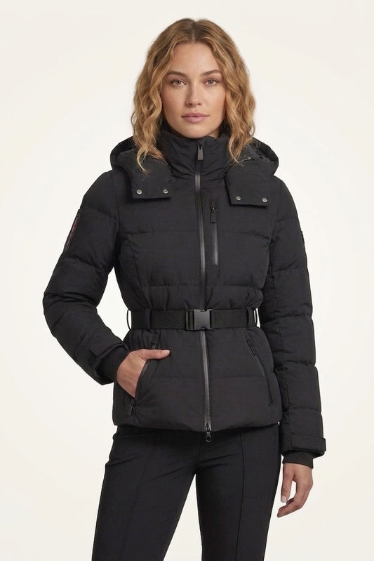 A woman modeling a black hooded puffer jacket with a front zipper, belt detail, and flap pockets, paired with black pants against a plain light background.