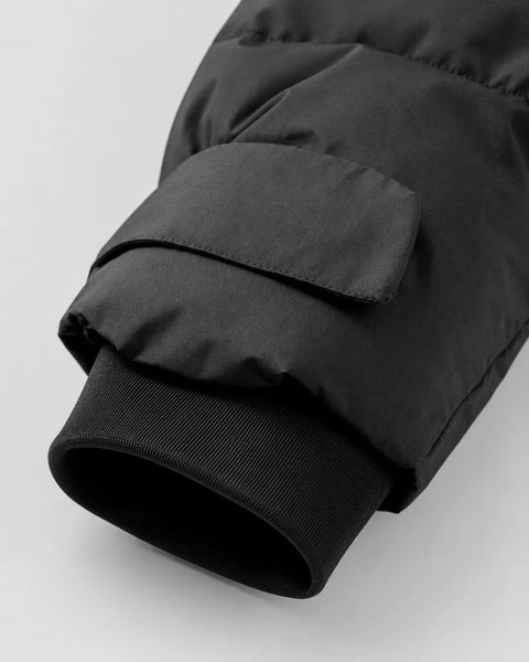 Close-up detail of a black puffer jacket sleeve showing the ribbed knit cuff and a flap pocket on the cuff, set against a light gray background.
