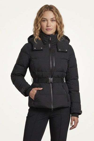 A woman modeling a black hooded puffer jacket with a front zipper, belt detail, and flap chest pockets, paired with black pants against a plain light background.