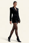 A model wearing a black velvet blazer, black lace tights, and black pointed-toe heeled ankle boots, holding a small black clutch against a solid light beige background.