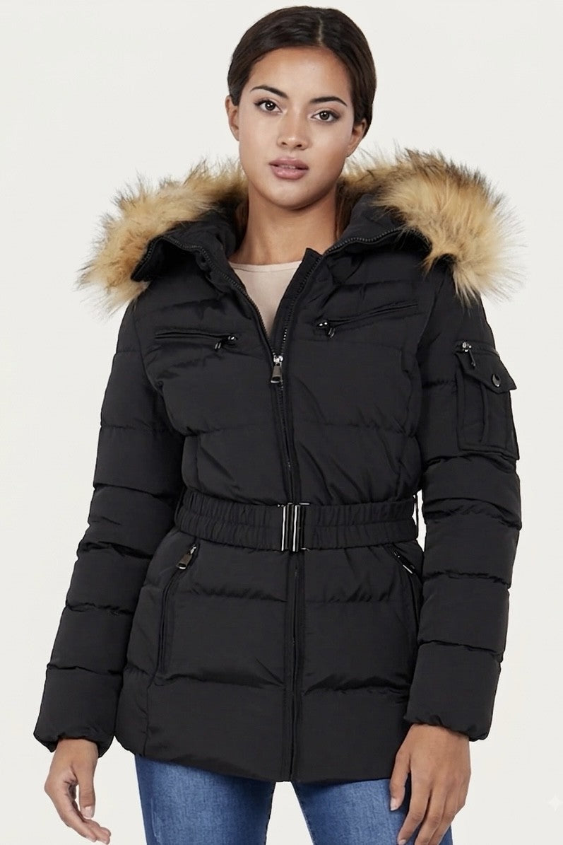 A woman wearing a black puffer parka jacket with a tan faux-fur trimmed hood and a matching black belt, paired with blue jeans, against a plain light background.-95