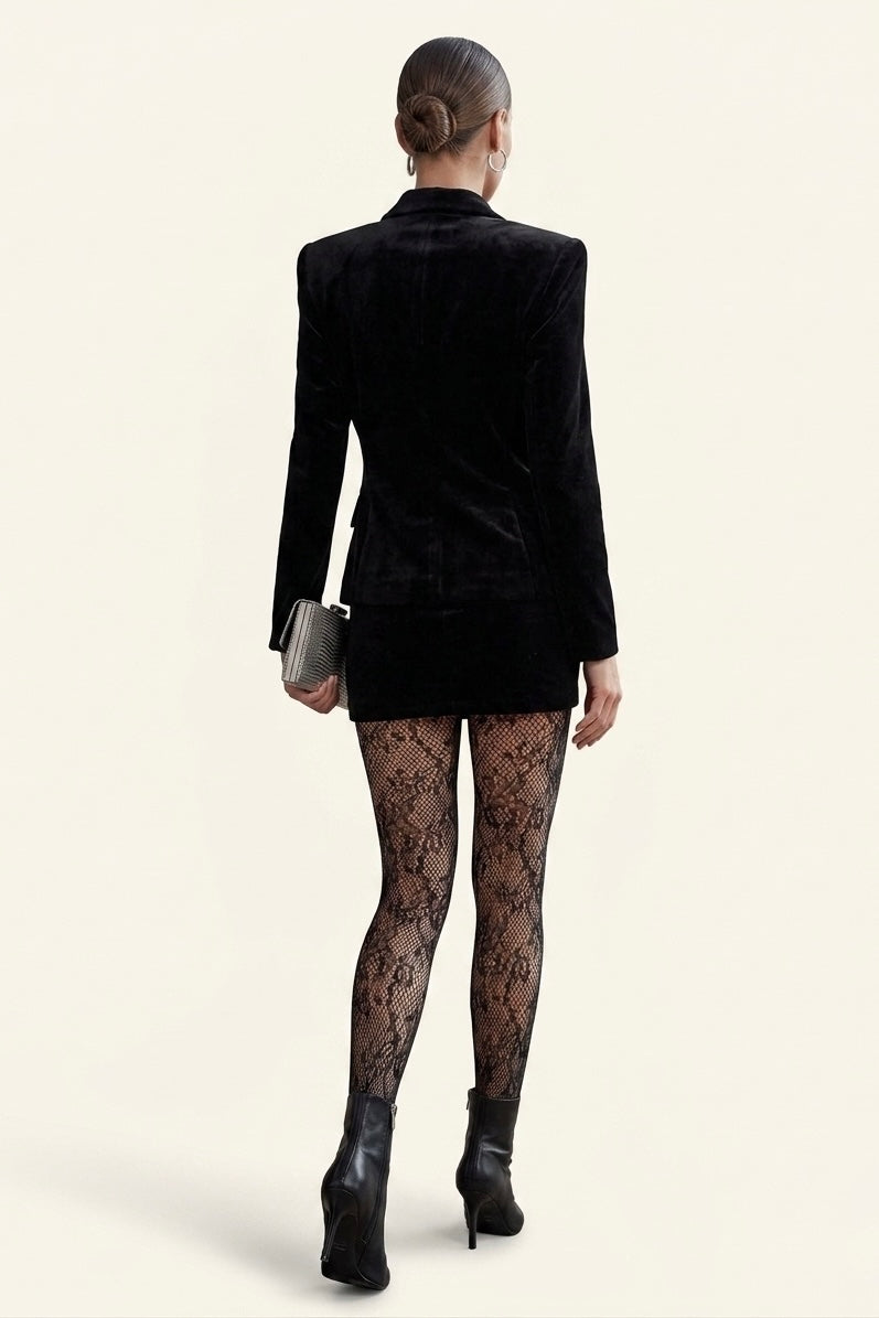 A model shown from the back wearing a black velvet blazer, a short black skirt, black lace tights, and black ankle boots, holding a small silver clutch against a light beige background.