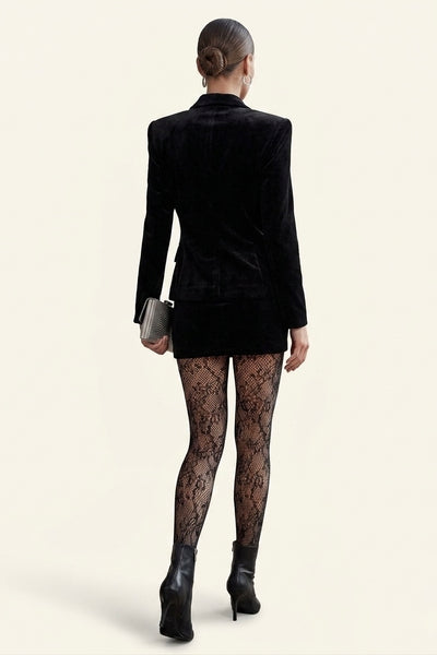 A model shown from the back wearing a black velvet blazer, a short black skirt, black lace tights, and black ankle boots, holding a small silver clutch against a light beige background.