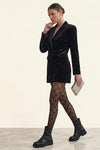 A woman models a black velvet blazer dress with a tie waist, paired with black fishnet tights and black leather ankle boots, holding a silver clutch against a warm beige background.