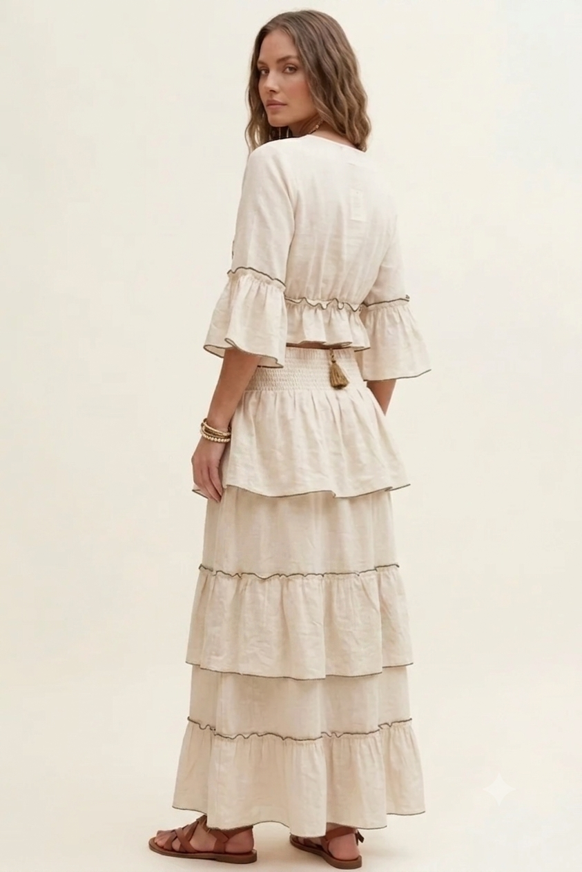 A woman wearing a cream-colored, tiered maxi dress with ruffled sleeves, shown from the back against a plain, warm off-white studio background.