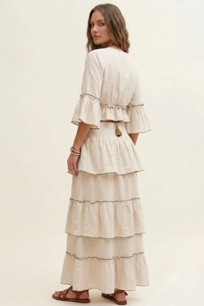 A woman wearing a cream-colored, tiered maxi dress with ruffled sleeves, shown from the back against a plain, warm off-white studio background.