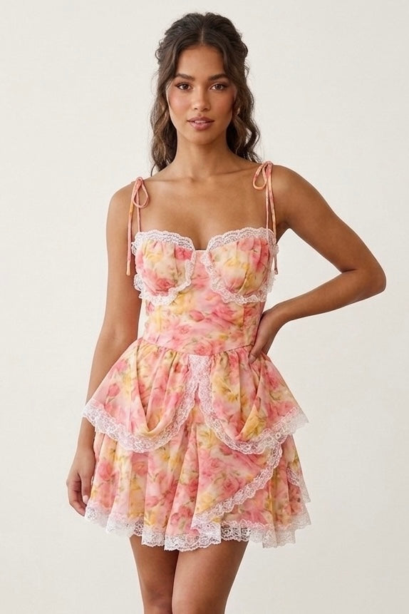 A woman modeling a pastel floral mini dress with a ruffled corset-style bodice, thin shoulder straps, and a layered ruffle skirt against a plain light background.
