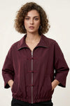 A woman with curly brown hair wearing a dark burgundy zip-up jacket with toggle closures, photographed against a plain light background.