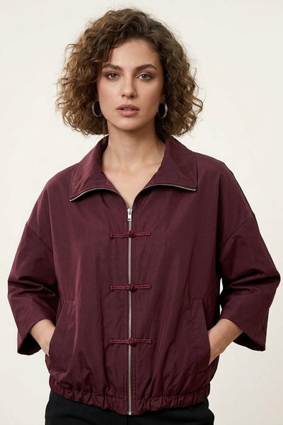 A woman with curly brown hair wearing a dark burgundy zip-up jacket with toggle closures, photographed against a plain light background.