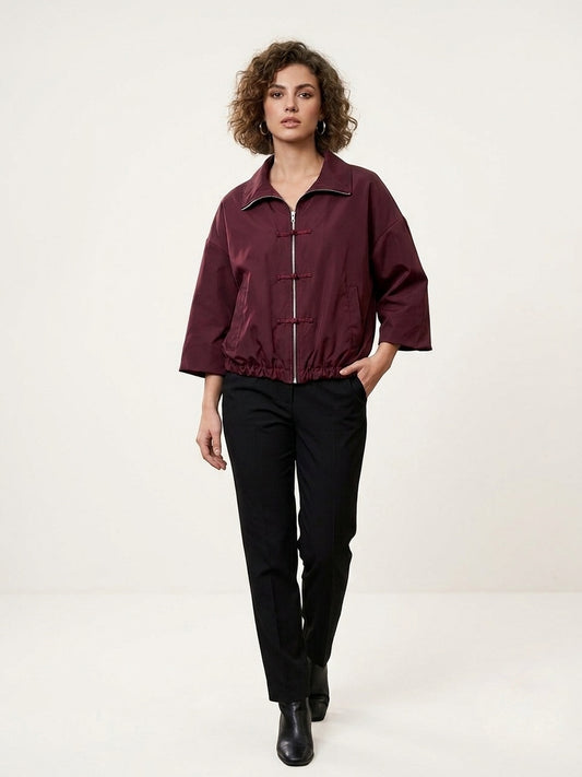 A woman modeling a cropped, oversized burgundy zip-up jacket with three-quarter sleeves and black trousers, standing against a plain off-white studio background.