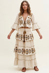A woman modeling a cream-colored bohemian-style two-piece set featuring a cropped jacket with bell sleeves and a matching tiered maxi skirt, both decorated with intricate brown and gold geometric embroidery.