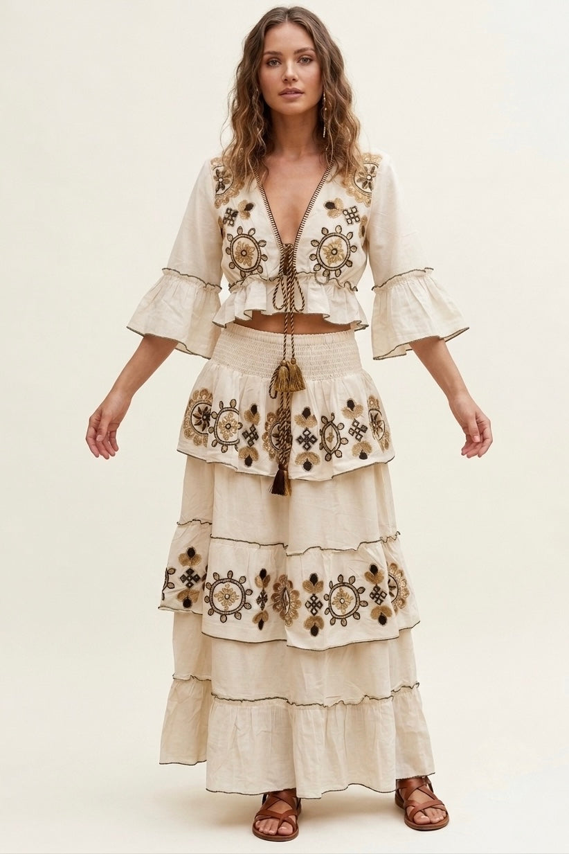 A woman modeling a cream-colored bohemian-style two-piece set featuring a cropped jacket with bell sleeves and a matching tiered maxi skirt, both decorated with intricate brown and gold geometric embroidery.