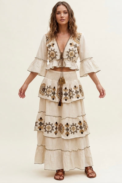 A woman modeling a cream-colored bohemian-style two-piece set featuring a cropped jacket with bell sleeves and a matching tiered maxi skirt, both decorated with intricate brown and gold geometric embroidery.