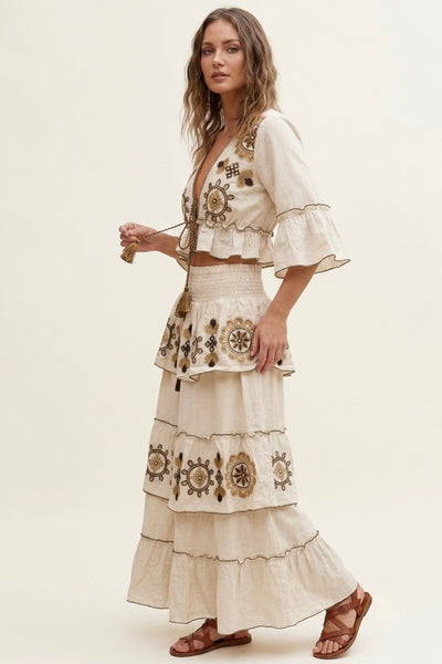 A woman modeling a bohemian-style two-piece outfit consisting of a cream-colored embroidered crop top with ruffled bell sleeves and a matching tiered maxi skirt with floral embroidery, paired with brown strappy sandals against a plain cream background.