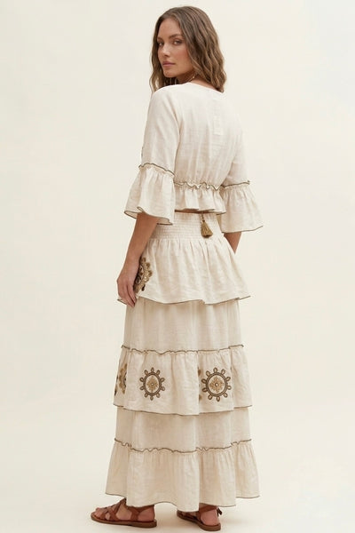 A woman modeling a cream-colored tiered maxi dress with embroidered details and ruffled sleeves, shown from a back-three-quarter view against a plain cream background.