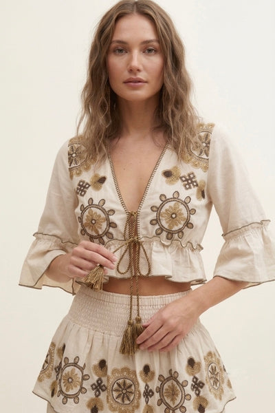 A woman modeling a bohemian-style two-piece outfit featuring a cream-colored crop top and matching skirt with detailed brown and gold geometric and floral patterns.