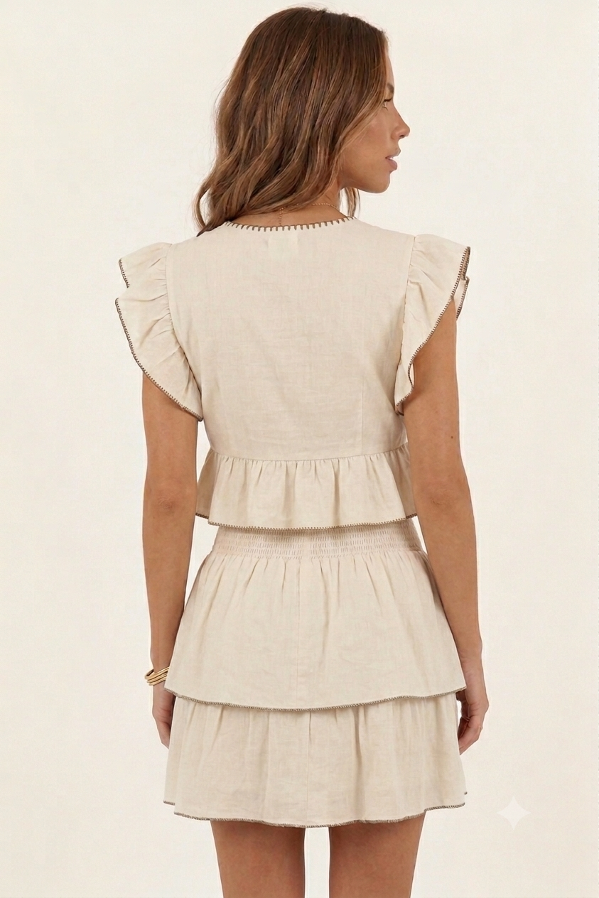 A woman shown from the back wearing a light beige linen-like dress with ruffled cap sleeves, a peplum waist, and a tiered skirt against a plain off-white background.