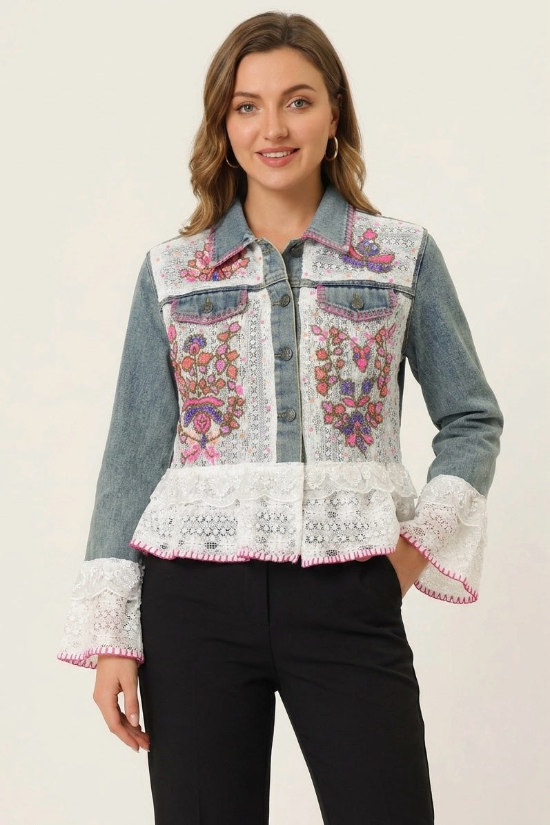 A woman modeling a denim jacket with intricate floral embroidery on the front, white lace applique on the collar, hem, and cuffs, and black pants, set against a plain light background.