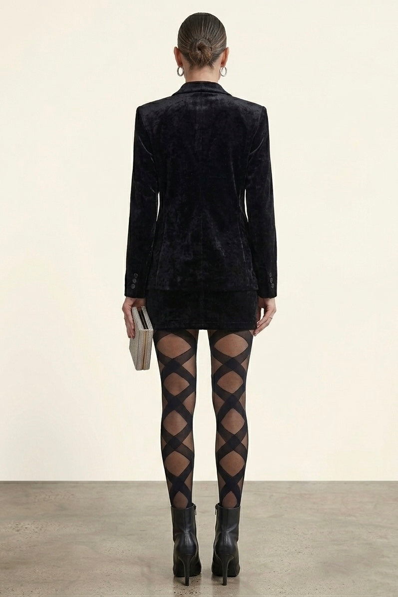 A model shown from the back wearing a black velvet blazer, a black mini skirt, black diamond-patterned tights, and black ankle boots, holding a small metallic clutch against a neutral studio backdrop.