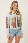 A woman modeling a light-wash denim jacket covered in colorful graphic prints and patchwork designs, paired with light blue denim shorts against a plain off-white studio background.