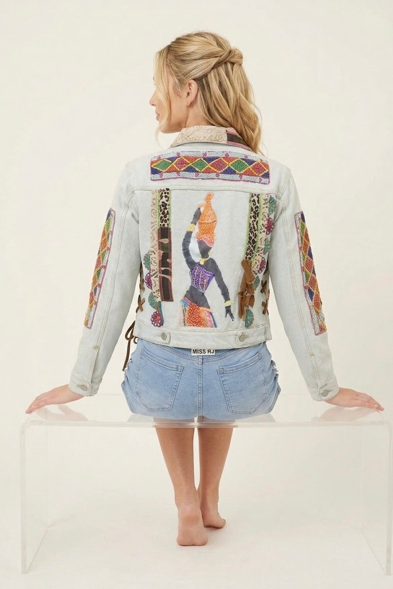 A woman stands barefoot on a clear acrylic bench, viewed from the back, wearing a light-wash denim jacket with colorful Egyptian-themed graphic prints and blue denim shorts against a plain off-white studio background.
