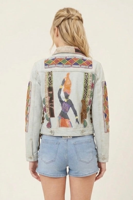 A woman shown from the back wearing a light-wash denim jacket with colorful Egyptian-themed patchwork embroidery and a large central figure design, paired with light blue denim shorts against a plain off-white background.