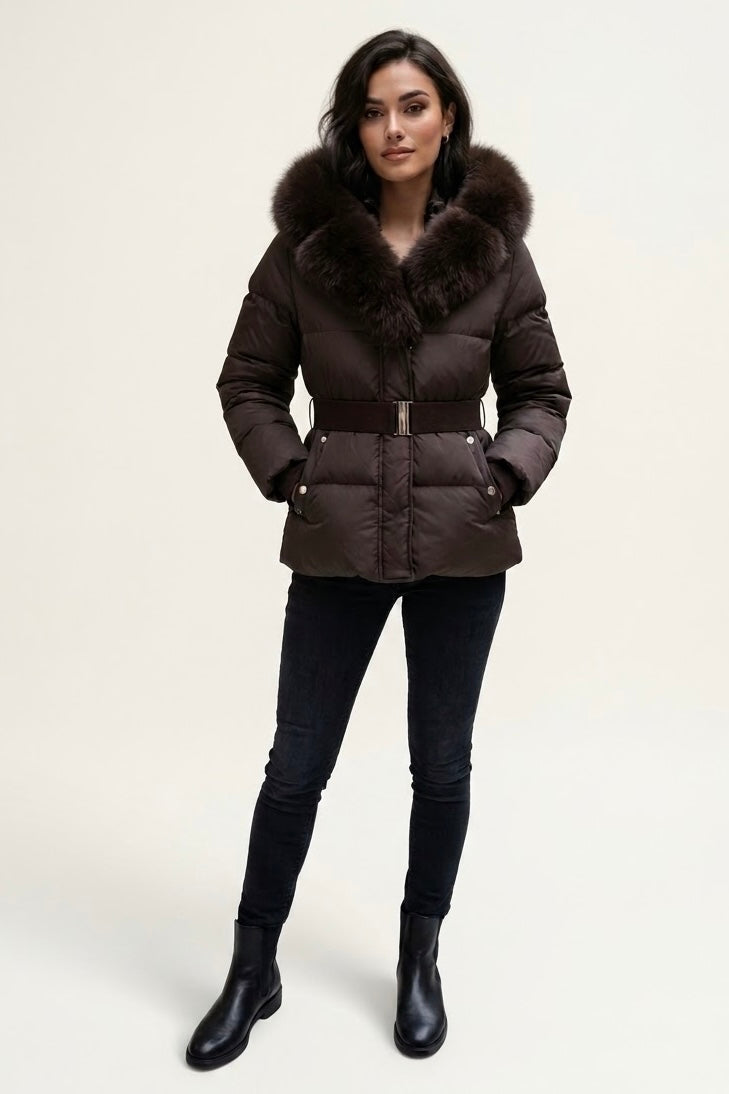 A woman modeling a dark brown puffer jacket with a large brown fur-trimmed collar, paired with black skinny jeans and black ankle boots, against a plain light beige studio background.