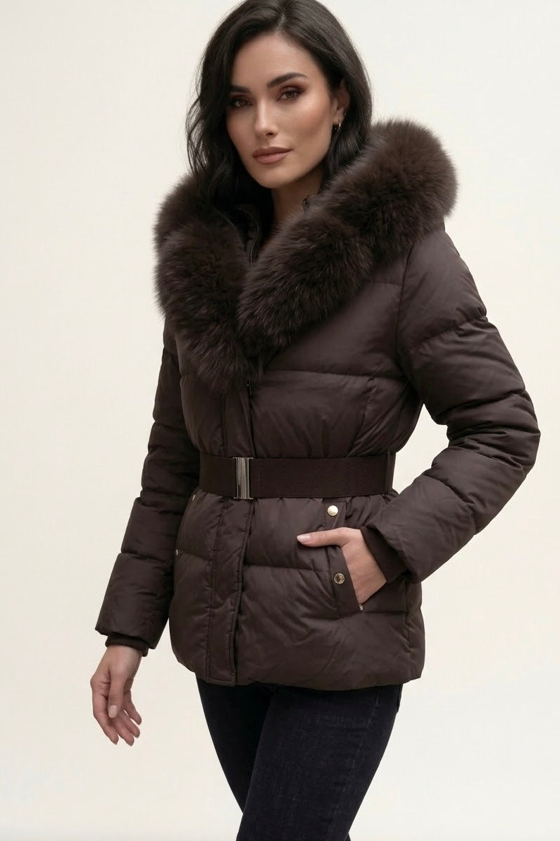 A woman wearing a dark brown puffer jacket with a large brown fur-trimmed collar and a matching belt, paired with dark jeans, against a plain light background.