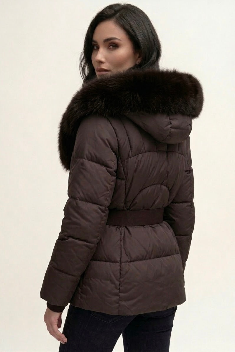 A woman wearing a dark brown puffer jacket with a large dark brown faux-fur trimmed hood and a matching belt, shown from the back against a plain off-white background.