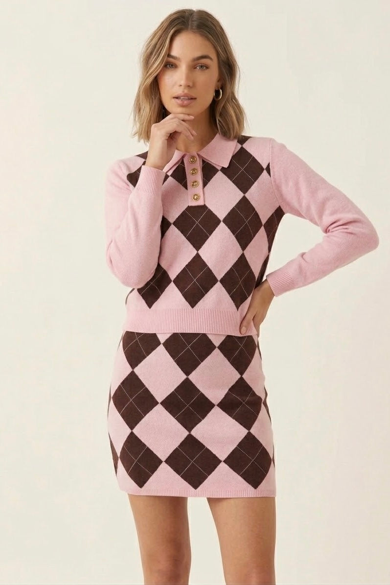 A woman modeling a pink long-sleeve polo-knit mini dress with a brown argyle pattern on the front and skirt, against a plain light beige background.