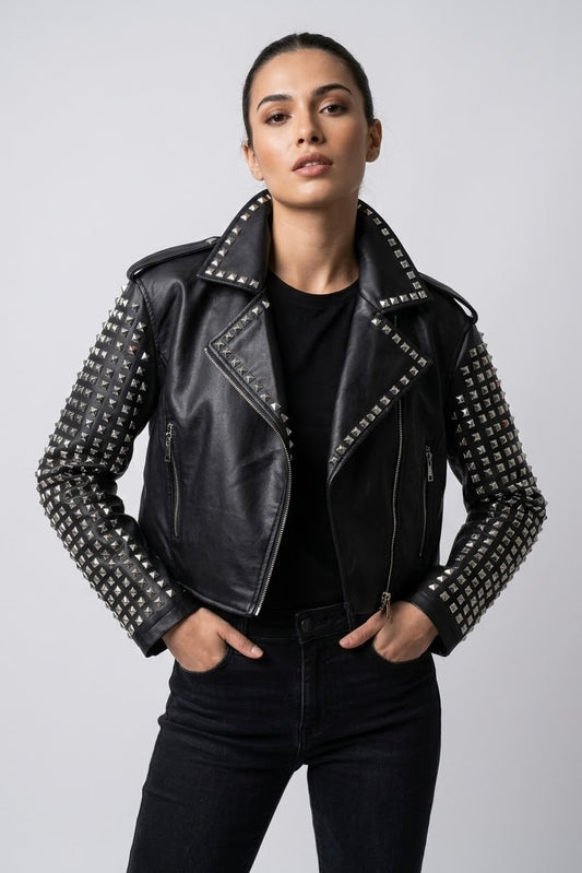 A woman modeling a black leather moto jacket with silver studs on the sleeves and collar, worn over a black top and dark jeans against a light gray studio background.