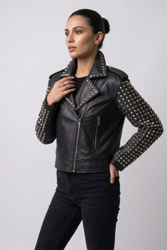 A woman modeling a black leather moto jacket with silver studs on the sleeves and shoulders, paired with black jeans, against a light gray studio background.