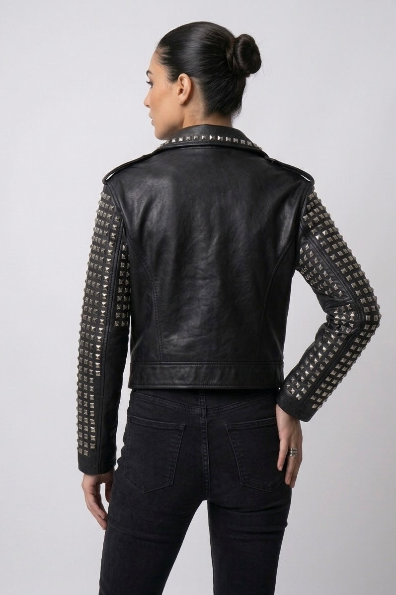 A woman shown from the back wearing a black leather jacket with silver studs on the sleeves, paired with black jeans, against a light gray studio background.