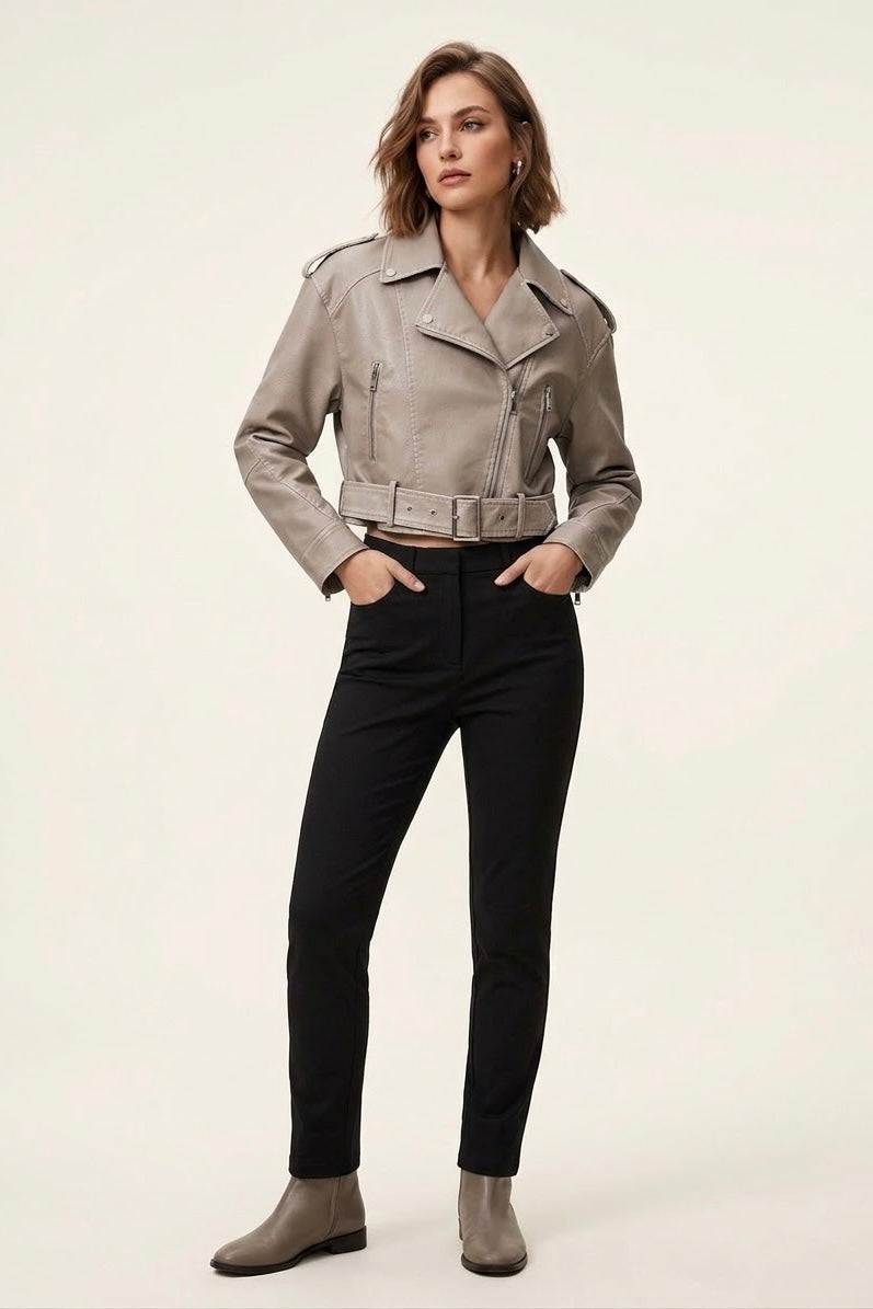 A woman modeling a cropped taupe faux-leather moto jacket with black pants and taupe ankle boots, standing against a plain off-white studio background.
