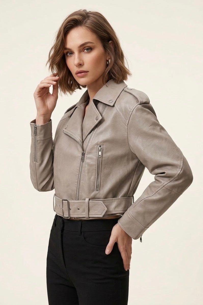 A woman modeling a cropped, light taupe faux-leather moto jacket with a matching belt, paired with black pants, against a solid off-white background.