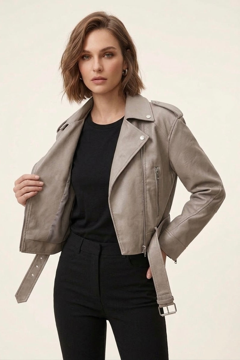 A woman modeling a taupe faux-leather moto jacket with a matching belt, worn over a black top and black pants against a light beige studio background.