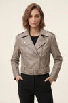 A woman modeling a taupe faux-leather moto jacket with silver hardware, worn over a black top and black pants, against a plain light beige background.