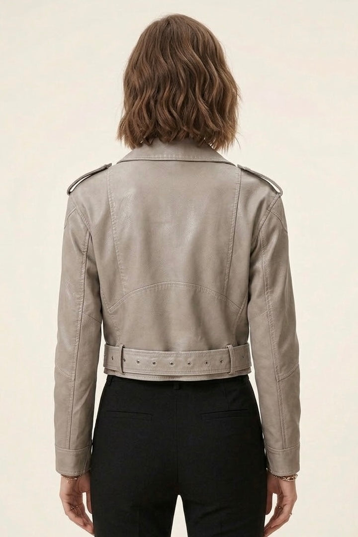 A woman viewed from the back wearing a cropped light taupe leather jacket with shoulder epaulets and a matching belt, paired with black pants, against a plain off-white background.