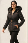 A woman wearing a black puffer jacket with a fur-trimmed hood, gold-tone zippers, and a matching belt, standing against a light textured wall.