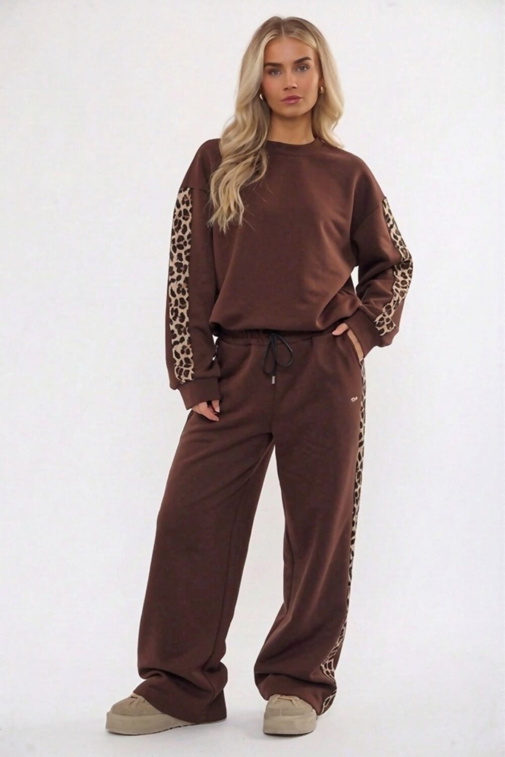 A woman modeling a brown two-piece lounge set featuring leopard-print sleeve and side stripe details, consisting of a crewneck sweatshirt and matching wide-leg sweatpants, set against a plain white studio background.