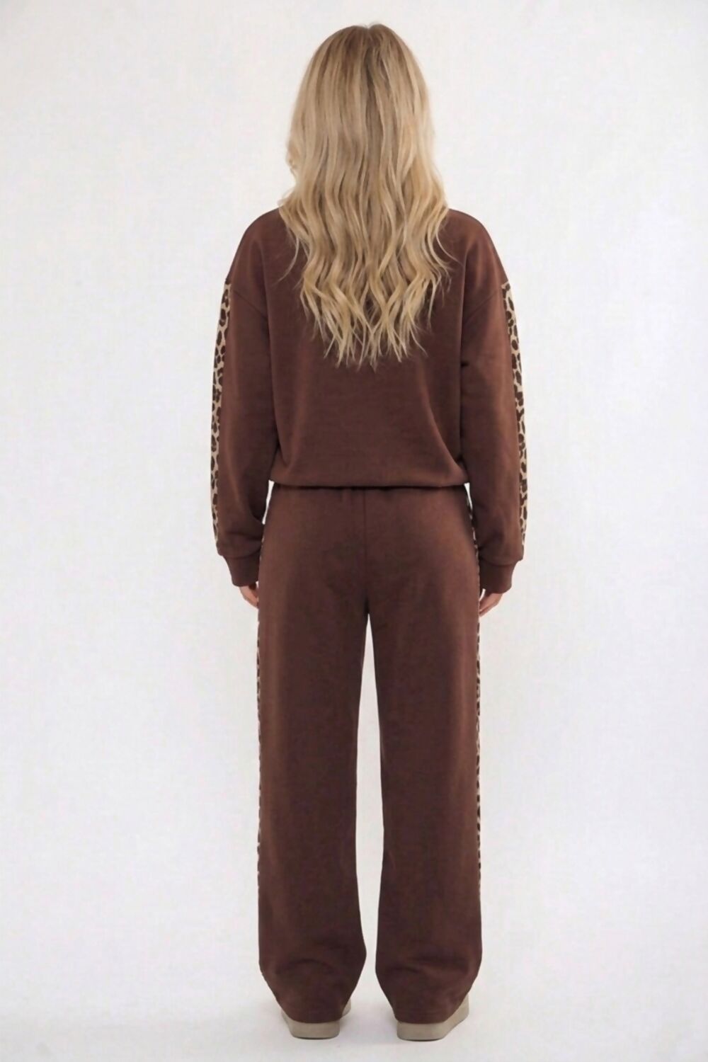 A woman viewed from behind wearing a brown two-piece lounge set with leopard-print sleeve and side details, against a plain white studio background.