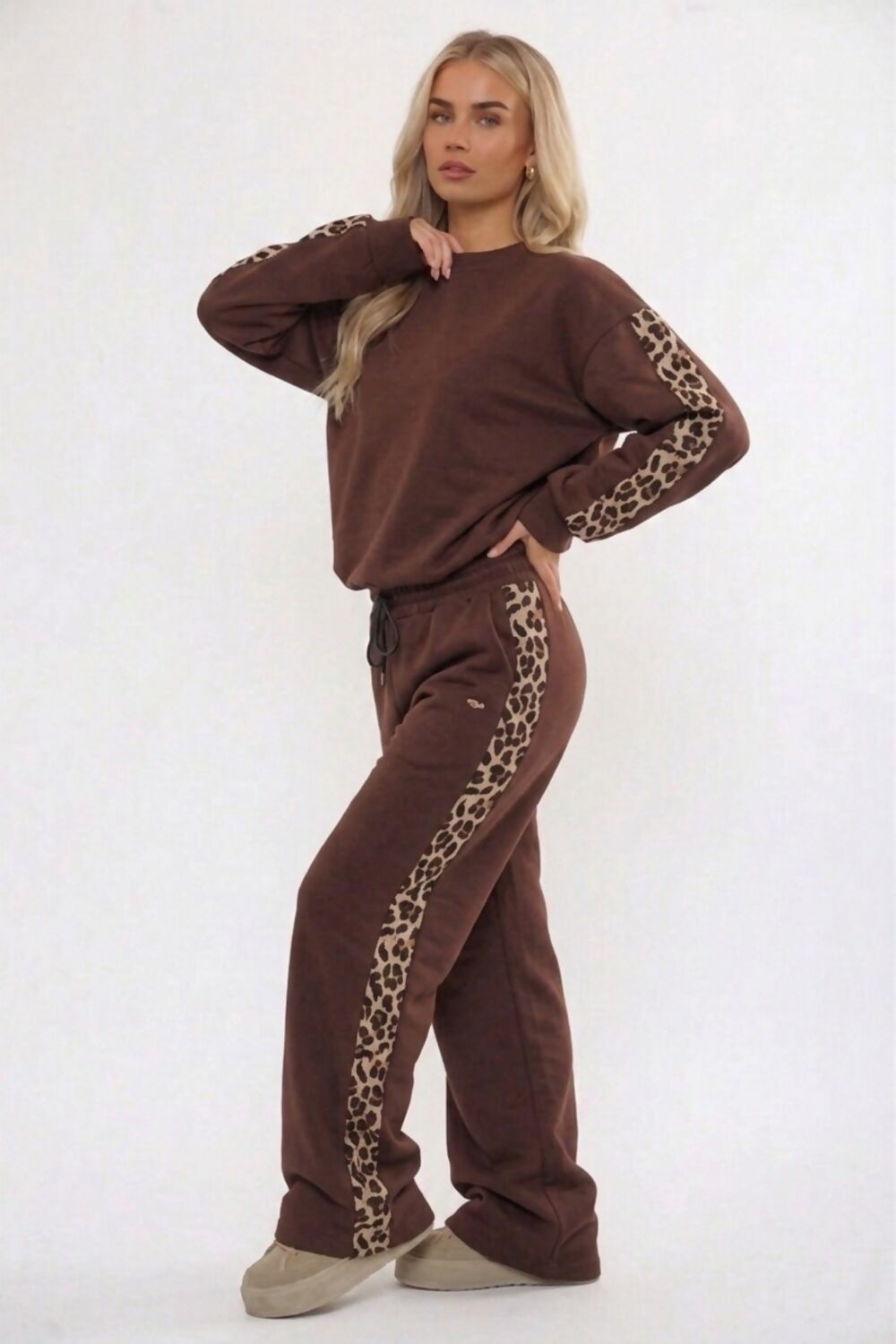 A woman modeling a matching brown lounge set with leopard print details, consisting of a long-sleeve sweatshirt and wide-leg sweatpants, against a plain white studio background.