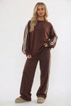 A woman modeling a matching brown lounge set consisting of a long-sleeve sweatshirt and wide-leg sweatpants, both featuring white stripes down the sleeves and legs, against a plain white studio background.