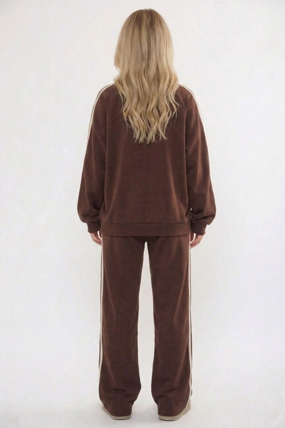 A woman viewed from behind wearing a matching brown sweatshirt and sweatpants set with white side stripes, against a plain white studio background.