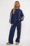 A woman modeling a navy blue sweatshirt and matching wide-leg sweatpants set, both featuring white stripes down the sleeves and legs, against a plain white studio background.