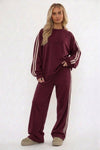 A woman modeling a matching brown lounge set consisting of a long-sleeve sweatshirt and wide-leg sweatpants, both featuring white stripes down the sleeves and legs, against a plain white studio background.